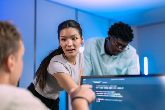 Woman leans in to talk with colleagues around a monitor of code during a focused problem-solving session in a blue-lit office.