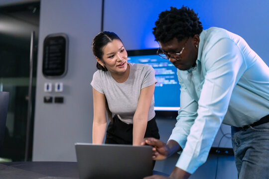 Woman and a colleague lean over a laptop to review code together in a blue-lit modern technology office with a screen behind them.