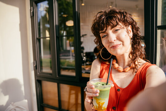 Stylish middle aged woman taking selfie with colorful iced matcha drink at modern urban cafe