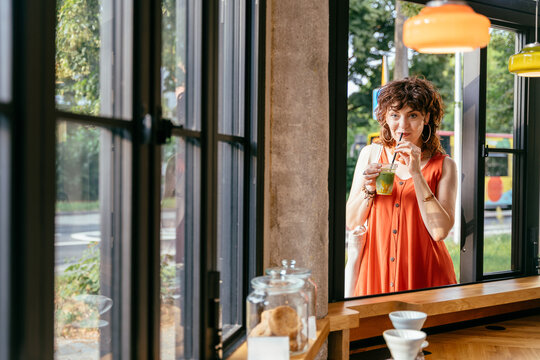 Stylish middle aged woman drinking iced matcha by open window at contemporary urban cafe