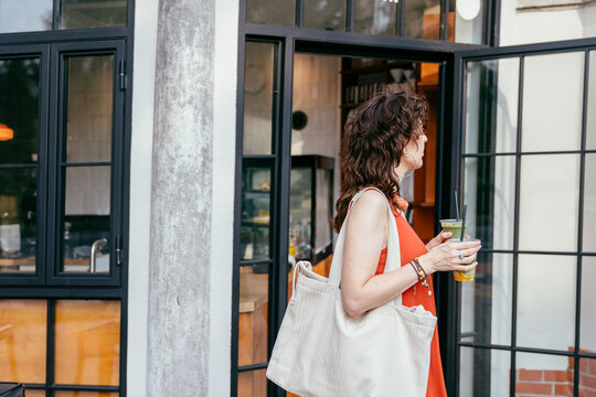 Stylish middle aged woman leaving modern cafe with iced matcha and reusable canvas tote bag