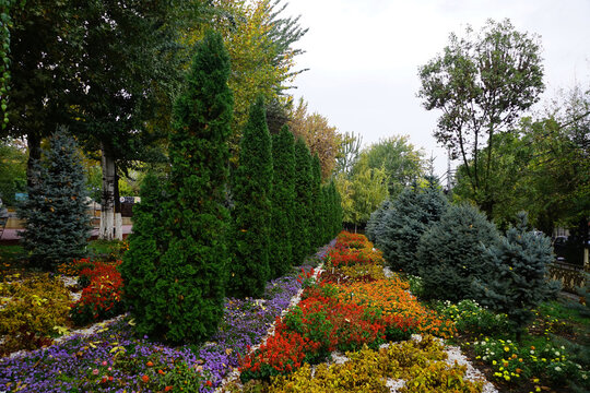 A neatly landscaped garden features colorful flowerbeds and manicured evergreen trees in a public park under an overcast sky in Osh, Kyrgyzstan