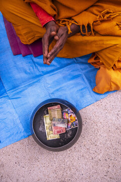 A monk praying with his hands clasped on a blue mat along the Kora pilgrimage route in Dharamsala, Himachal Pradesh, India. A bowl of offerings contains Indian rupee bills.