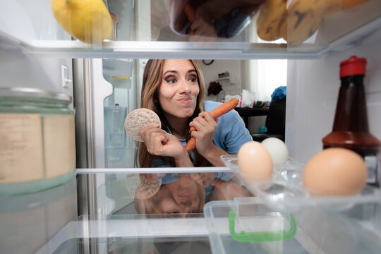 Hesitant Woman Holding Rice Cake And Carrots Near Open Refrigerator. Doubtful female looks inside fridge deciding between healthy snacks in kitchen interior