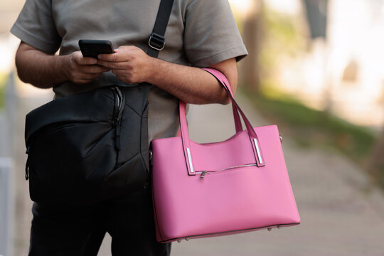 Helpful Husband Holding Bright Pink Handbag While Using Smartphone. Man carries his wife purse outdoor checking mobile phone on city street 
