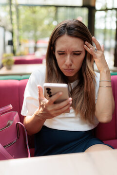 Squinting Young Woman Reading Text Message On Smartphone Inside Restaurant. Confused female strains her eyes looking at mobile screen upon terrace couch