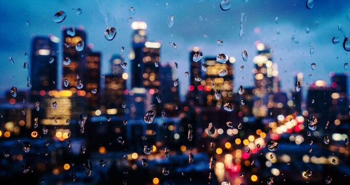 A moody cityscape of downtown Los Angeles viewed through a rain-soaked window, showcasing lights blurred by rain droplets and a dusk atmosphere.