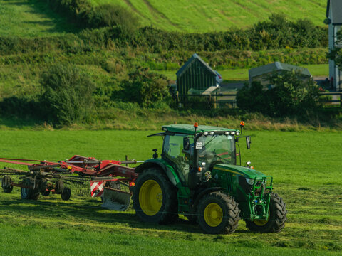 West Cork, Ireland, August 25, 2023. John Deere tractor and hay tedder working in a green pasture. Agricultural machinery.