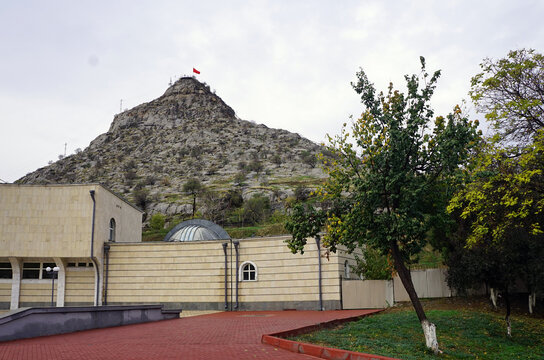 The sacred Sulaiman-Too Mountain, topped with a Kyrgyz flag, rises behind a modern stone complex under an overcast sky in Osh, Kyrgyzstan