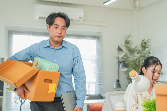 A serious man carries a cardboard box of belongings and his laptop as he prepares to leave the office, a distressed female colleague visible behind him  a classic depiction of layoff and job terminat
