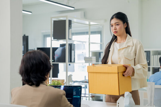 A woman sadly detaches her company phone from its lanyard strap while a colleague looks on, boxes of belongings nearby a quiet moment of returning corporate property during an office departure.