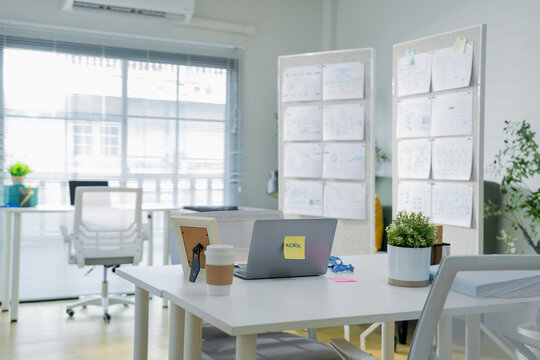 An empty white desk holds a closed laptop with a work sticky note, coffee cup, and picture frame, set against a bright office backdrop with wall mounted planning charts a clean, minimalist workspace s