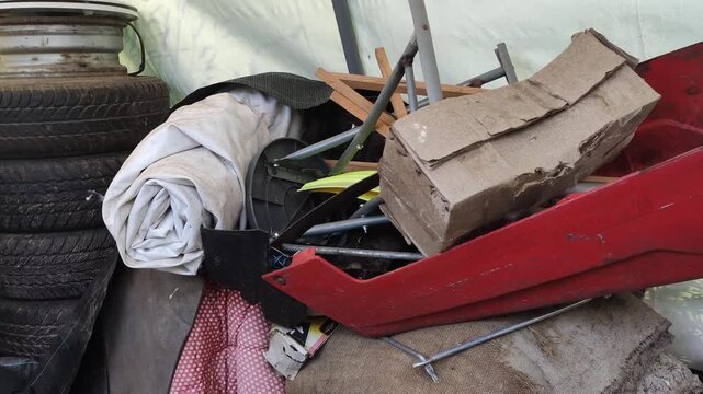 A jumble of miscellaneous discarded items, including a red plastic casing, fabric materials, bags, tools, and rods. Household waste, clutter, recycling, disposal, and environmental pollution concepts.