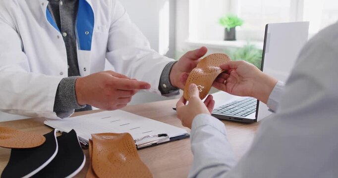 Orthopedist doctor shows orthopedic insoles to patient in medical clinic. They discuss custom orthotics and arch support to relieve foot pain. Concept medicine, foot healthcare and comfort.