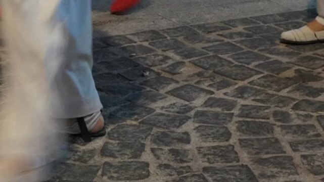 Feet wearing white shoes and trousers step briskly across dark cobblestones during an evening stroll. Such dynamic movement captures the lively rhythm of urban pedestrian life and city exploration.