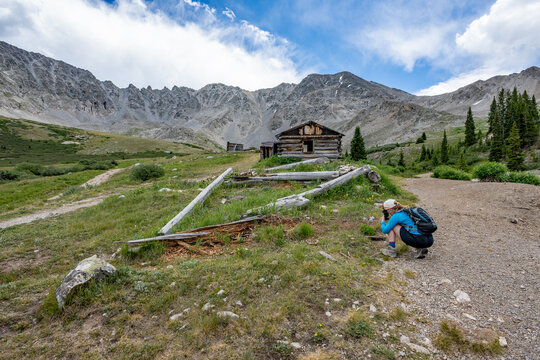 Woman hiker exploring and photographing abandoned mining cabins on Mayflower Gulch trail near Copper Mountain, Colorado under summer afternoon clouds.