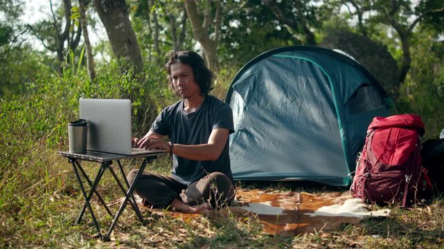 Remote worker using a laptop beside a camping tent in a grassy forest meadow, capturing work from nature, digital nomad lifestyle, outdoor workspace, and remote work.