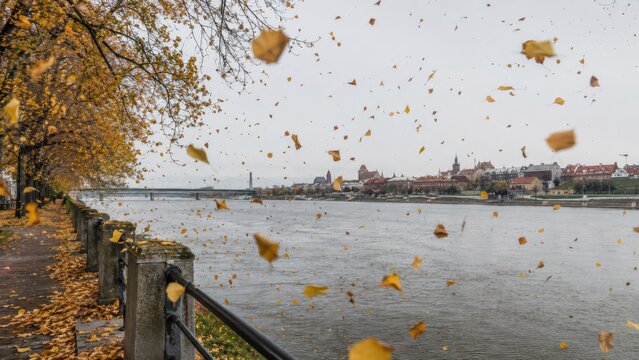 Herbststurm an einer Flusspromenade in Polen: Fallende Bl&auml;tter wirbeln &uuml;ber das Ufer und schaffen eine dynamische Stadtszene &ndash; atmosph&auml;risches Reisebild f&uuml;r Wetter und Saison.