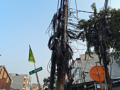 A chaotic and tangled web of overhead electrical cables and communication wires crisscrossing on utility poles in an urban street, showing urban infrastructure issues.