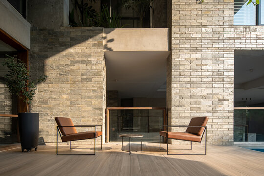 Modern Outdoor Terrace with Two Brown Lounge Chairs, Stone Walls, Glass Doors, and Natural Sunlight