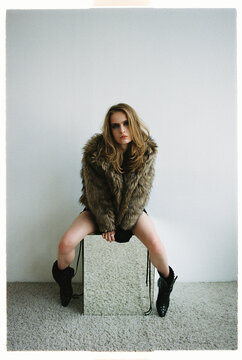 Model poses in a fur coat while sitting on a  mirror block