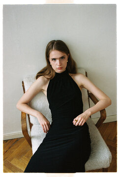 Model sits on chair wearing black dress in indoor setting