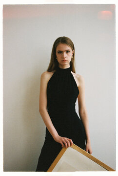 Model stands in studio holding a frame while wearing a black dress