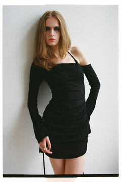 Model poses in black dress against a plain wall in a studio