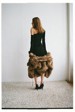 Woman stands in a simple room wearing a black dress and fur coat