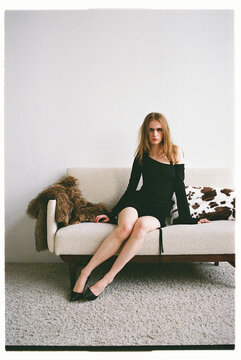 Model sits on a couch in a room with simple decor while wearing dress