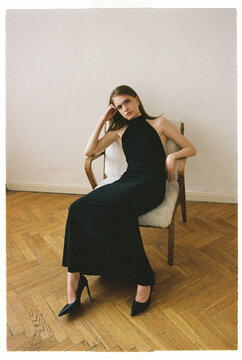 Woman sits in chair wearing black dress and high heels
