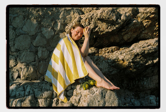 A woman dries her hair on sun-warmed coastal rocks