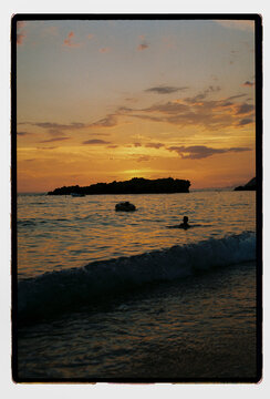 A person drifts through sunset-lit waves toward a rocky isle