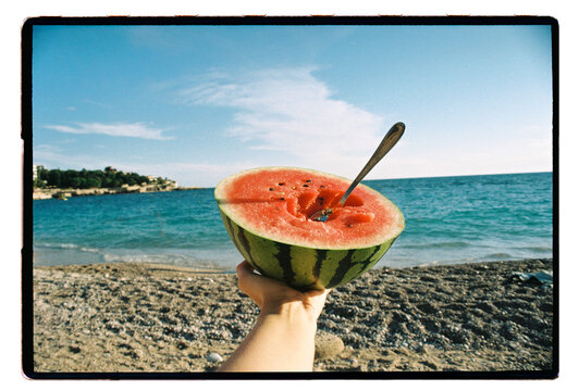 A hand holds half a watermelon over a pebble beach