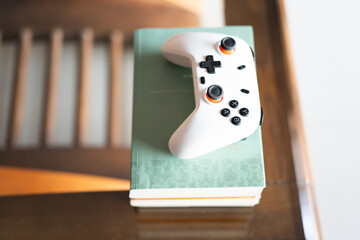 White gaming controller resting on stacked books atop wooden table in cozy indoor setting © chuangxin