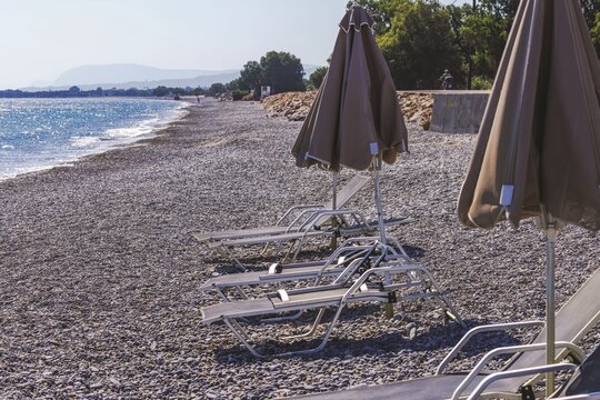 Close-up view of empty sun loungers and closed umbrellas on pebble beach beside sea. Crete. Greece.