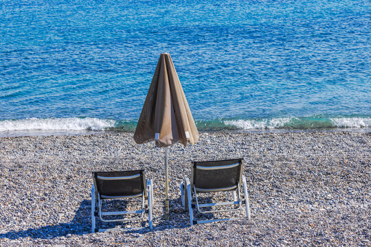 Beautiful view of two sun loungers and closed umbrella on pebble beach beside sea. Crete. Greece.