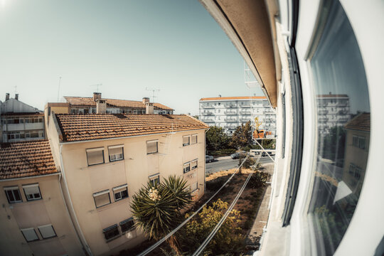Fisheye view from an apartment window overlooks a quiet residential district with terracotta rooftops, balconies, antennas, trees and sunlit communal streets