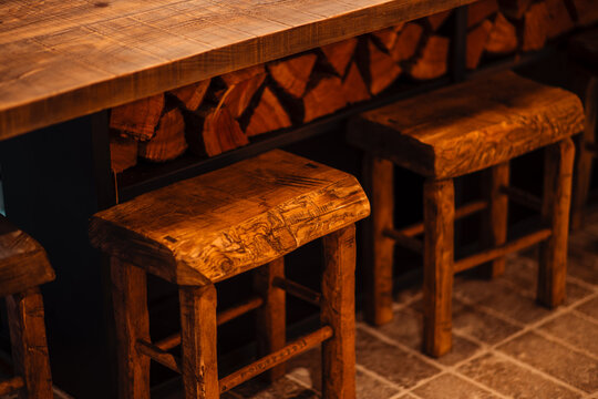Handcrafted timber stools line a thick wooden counter above neatly stacked firewood, revealing warm amber lighting, rich grain and cozy rustic hospitality decor