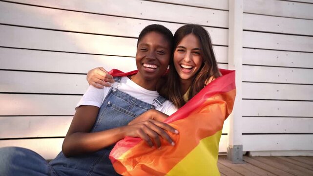 Happy multiracial lesbian couple looking at camera while hugging each other with a rainbow flag, celebrating Pride day. LGBTQIA concept.