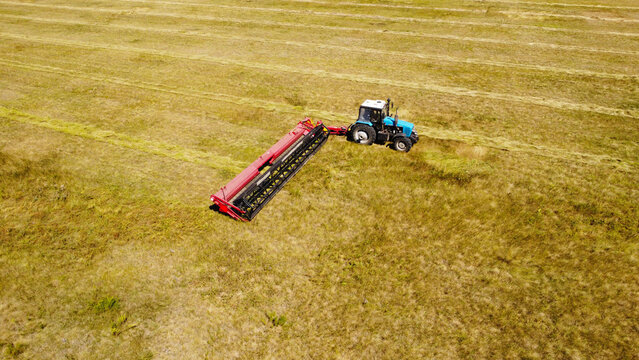 A high-angle aerial photograph capturing a blue tractor pulling a red trailing mower swather across a wide agricultural field