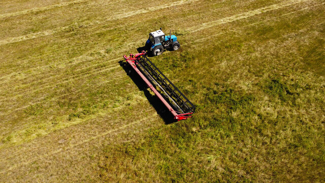 A high-angle aerial photograph capturing a blue tractor pulling a red trailing mower swather across a wide agricultural field