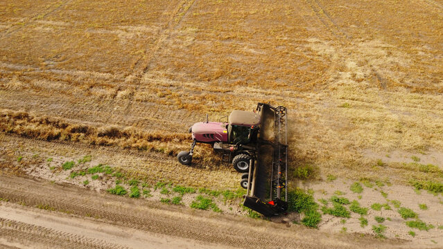 An aerial drone photograph capturing a self-propelled windrower swather operating across a vast, golden agricultural field