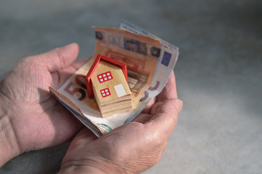 Elderly hands holding bills and a miniature house, concept for buying or selling a home, savings, insurance, donation for the homeless, mortgage and real estate crisis, copy space