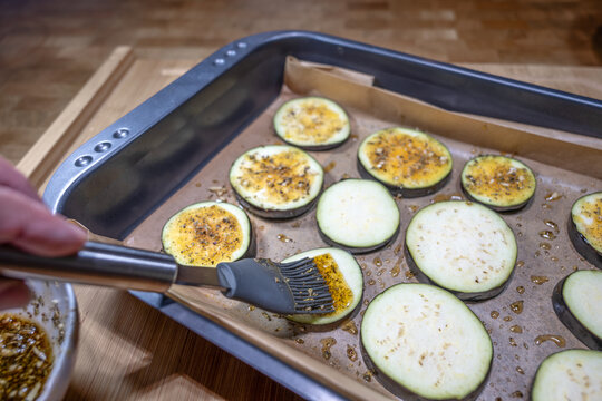 Eggplant slices on a baking sheet are brushed with seasoned oil using a silicone brush, preparing a flavorful vegetarian meal, selected focus