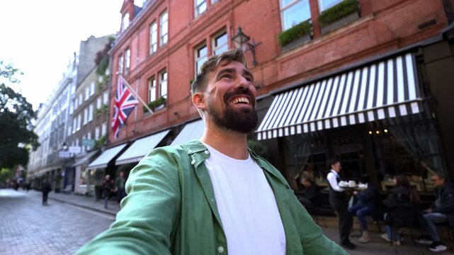 A happy caucasian traveler holds his smartphone to make a video call outdoors in the city. He is waving his hand and smiling directly at the camera with urban buildings in the background.