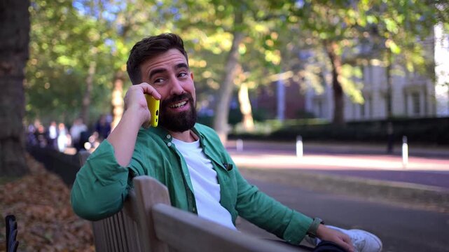Smiling Caucasian millennial man talking on his smartphone while sitting on a bench in an urban park. Casual outdoor scene showing mobile communication, relaxation and everyday life.