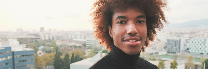 Fototapeta premium Close-up of young smiling guy posing at the camera while standing on the balcony modern city on the background. Panoramic