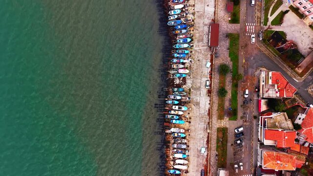 Drone view of moored boat beside coastal street, Bulgaria. Small fishing craft line a concrete shoreline near parked car and red roofed waterfront building.