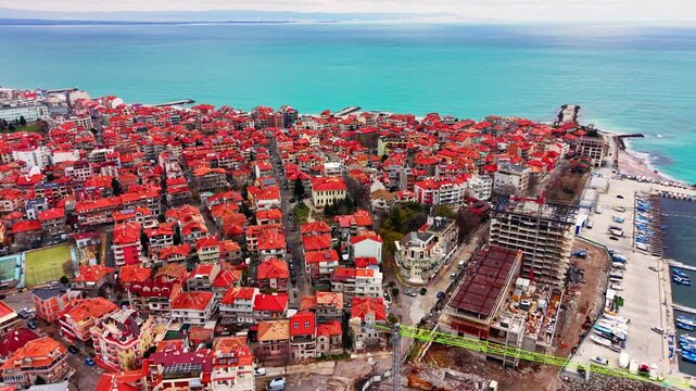 Elevated view of dense red roofed coastal town, Bulgaria. Row of terracotta rooftop stretch toward a turquoise bay beside a beachfront construction site.